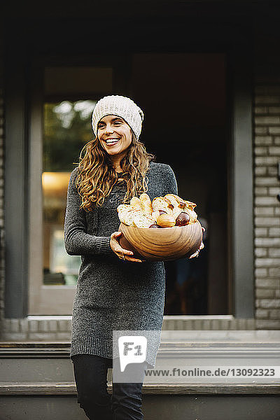 Happy woman carrying various breads in wooden bowl while standing against house entrance