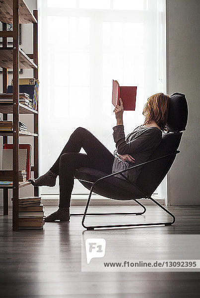 Frau  die zu Hause ein Buch hält und sich auf einem Stuhl am Fenster entspannt