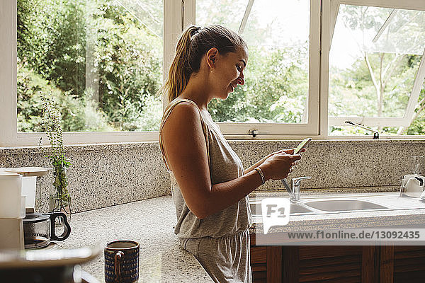 Side view of smiling woman using smart phone while standing in kitchen