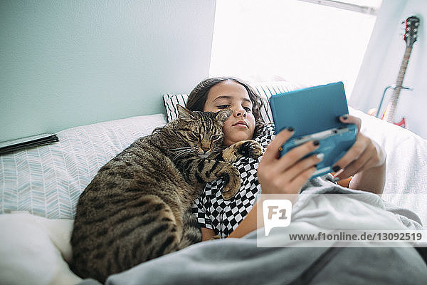 High angle view of girl playing video game while lying with cat on bed at home