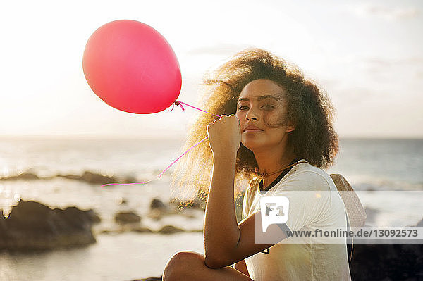 Portrait of teenage girl holding balloon while sitting on shore against sky