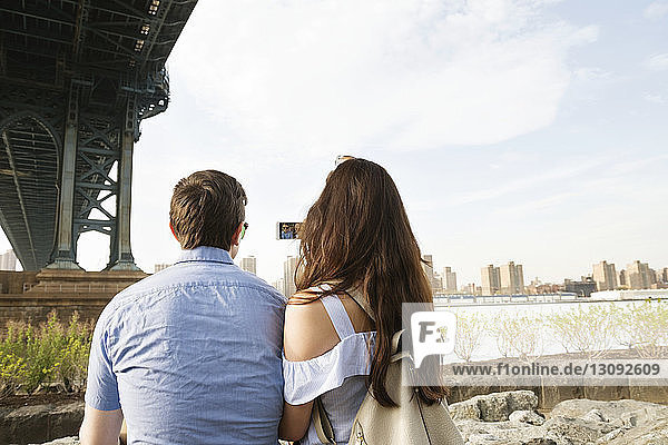Rear view of couple using phone while sitting below Manhattan bridge against sky
