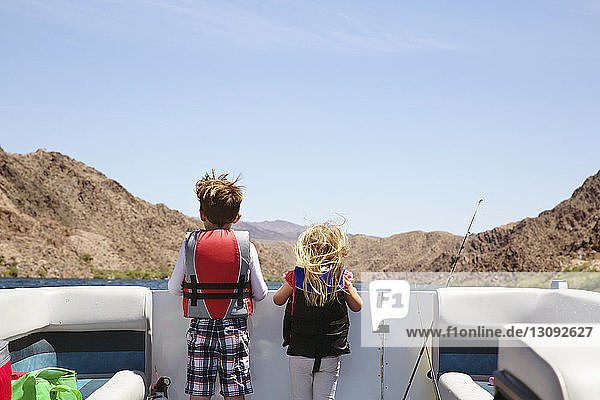 Rear view of siblings looking at mountains while standing in boat against blue sky