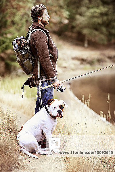 Seitenansicht eines Reisenden beim Angeln  während er mit seinem Hund am Seeufer sitzt