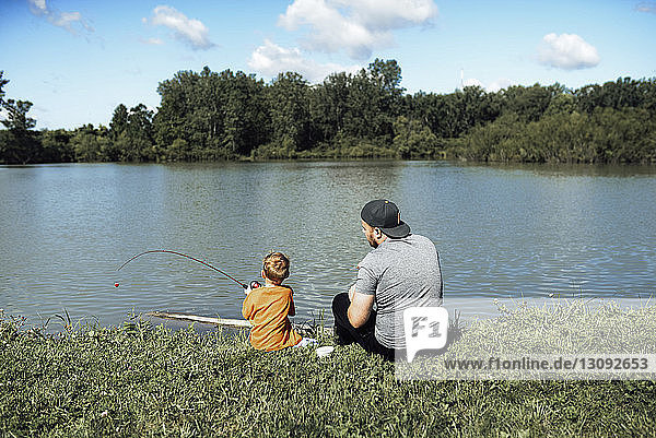 Rear view of son fishing while sitting with father at lakeshore against sky
