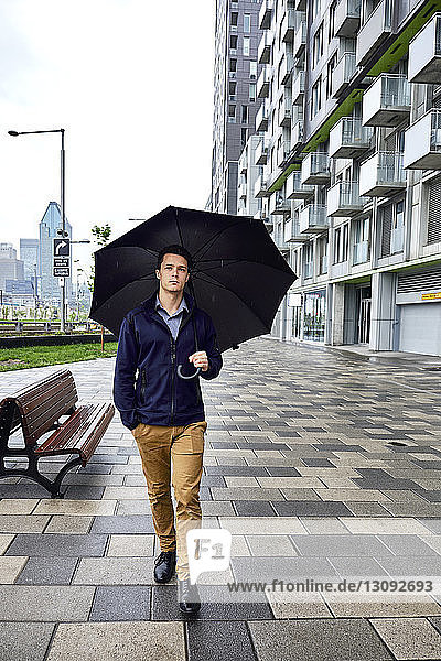 Full length of young businessman with umbrella walking on footpath in city during rainfall