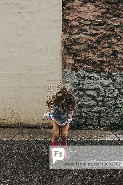 Verspieltes Mädchen springt in Pfütze auf Straße gegen Mauer