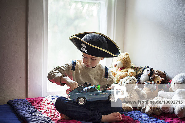 Boy wearing pirate hat playing with toy car while sitting on bed at home