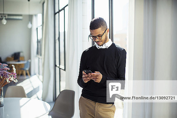 Businessman using mobile phone while standing by window at office