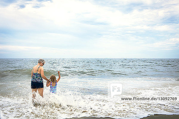 Rear view of granddaughter and grandmother holding hands while walking on shore at beach