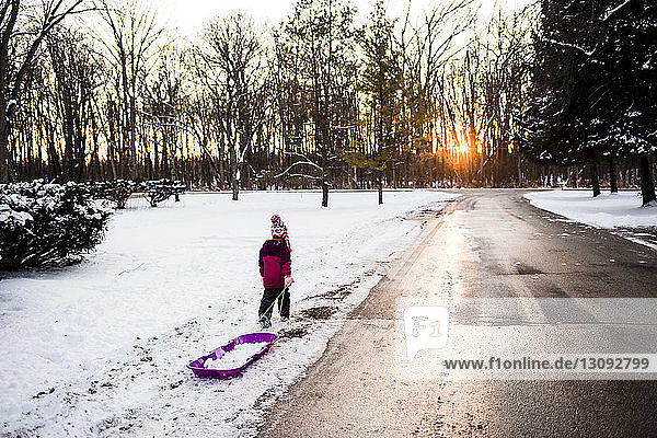 Rear view of girl pulling sled while walking on snow by road during winter at sunset