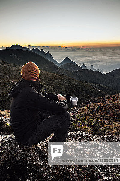 Seitenansicht eines Mannes  der einen Becher hält  während er bei Sonnenuntergang auf einem Berg sitzt