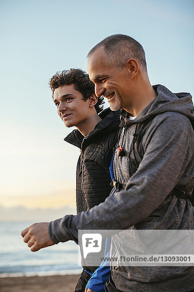 Side view of happy father and son walking at beach against sky