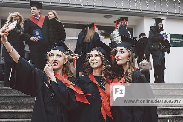 Happy female friends wearing graduation gowns taking selfie while standing on steps