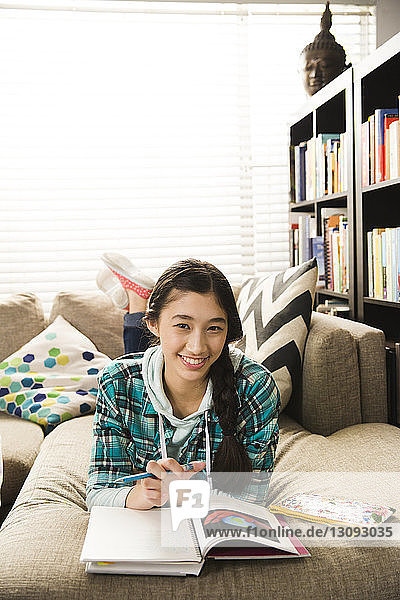 Portrait of confident teenage girl doing homework while lying on couch at home