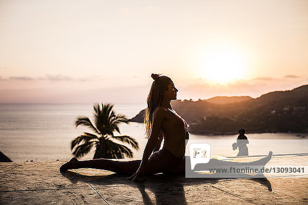 Woman practicing yoga on retaining wall against sea and sky during sunset