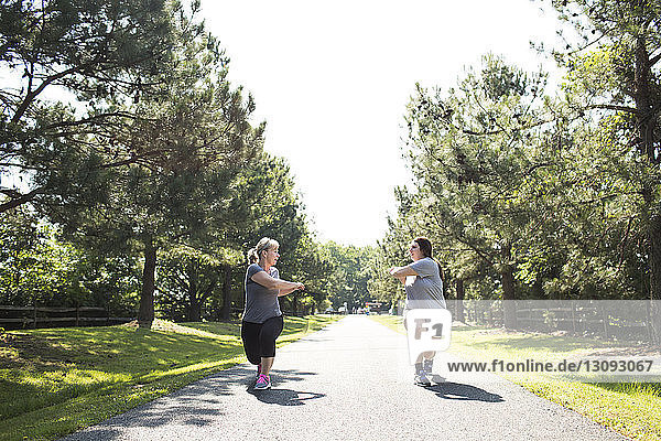 Mother and daughter stretching arms and legs on footpath at park during sunny day