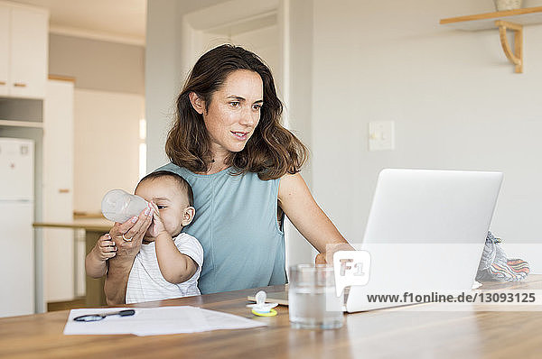 Frau füttert Sohn  während sie am Tisch einen Laptop benutzt