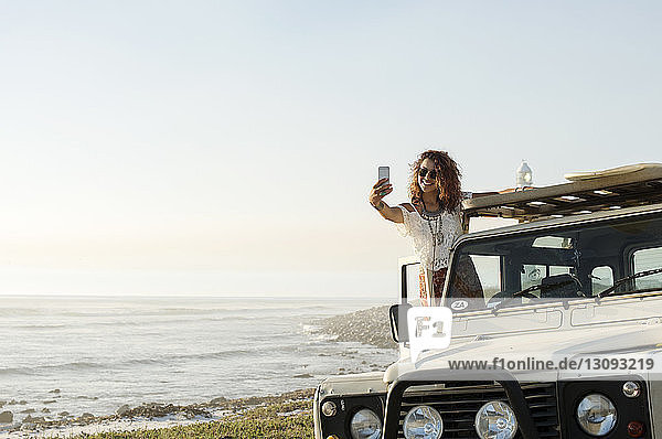 Happy woman taking selfie while standing on off-road vehicle at beach