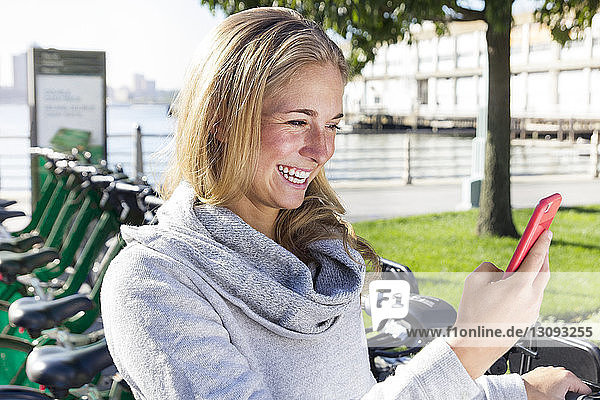Cheerful woman using phone while standing against bicycle rack in city