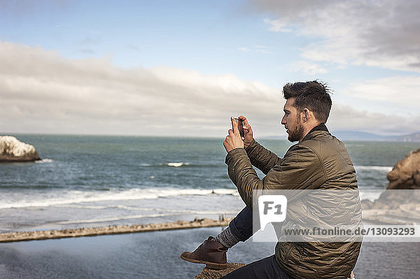 Side view of man photographing sea through smart phone against sky