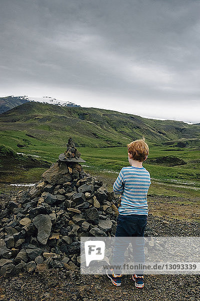 Boy standing by heap of rocks on field against cloudy sky