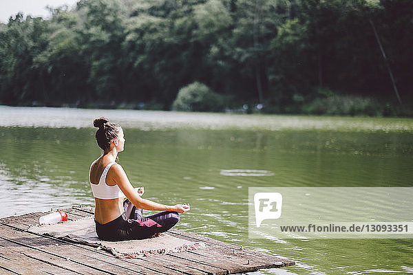 Side view of woman meditating while sitting on pier over lake in forest