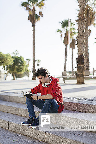 Serious teenage boy reading book while sitting on steps in city