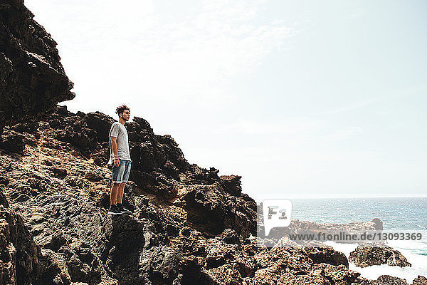 Side view of young man standing on rocks by sea against sky