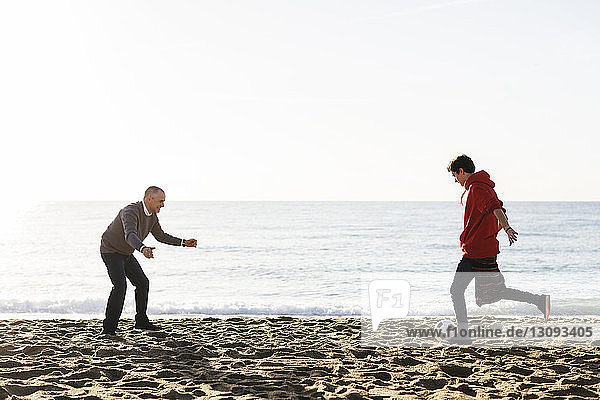 Son kicking soccer ball while father defending at beach against clear sky