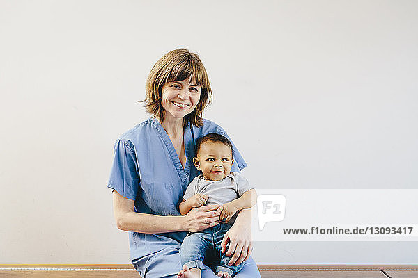 Porträt einer lächelnden Ärztin  die einen kleinen Jungen hält  während sie auf einer Bank vor weissem Hintergrund sitzt
