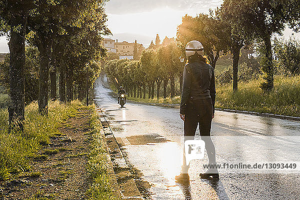 Rückansicht einer Frau  die steht  während ihr Freund bei Sonnenuntergang auf nasser Straße Motorrad fährt