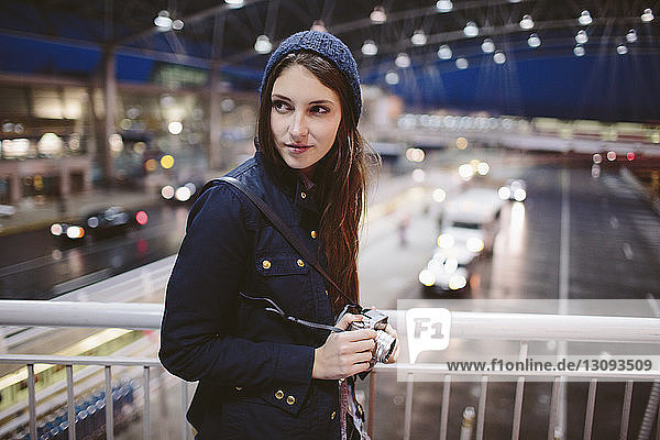 Woman holding camera looking away while leaning on railing in airport