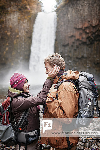 Happy female hiker touching man's face while standing against waterfall