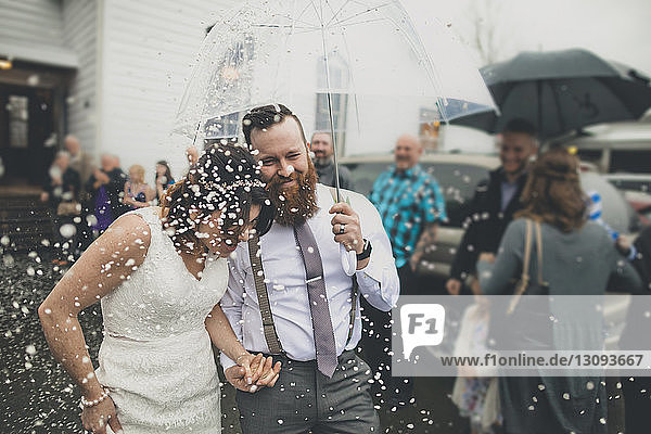 Glückliches frisch verheiratetes Paar geht mit einem Regenschirm spazieren,  während die Menschen während der Hochzeitszeremonie im Hintergrund stehen