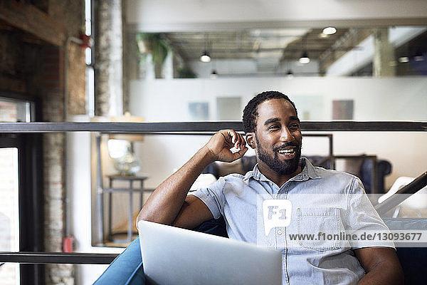 Glücklicher Geschäftsmann mit Laptop schaut weg  während er im Büro auf dem Sofa sitzt