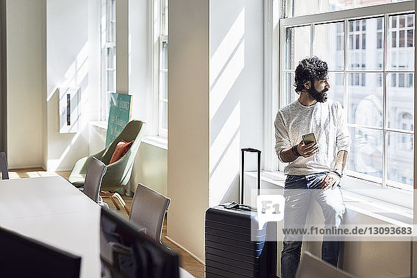Businessman looking through window while holding smart phone in creative office