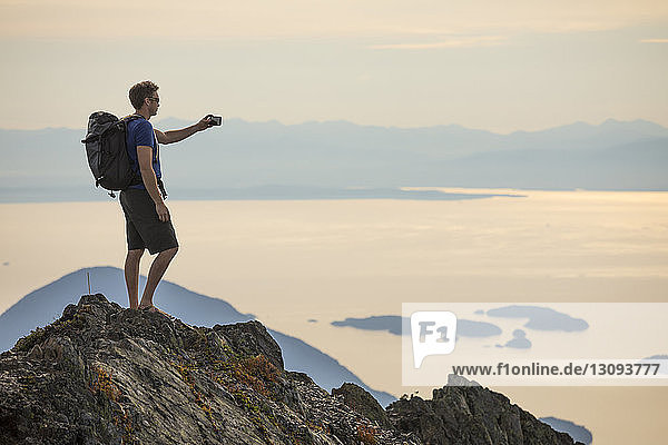 Side view of hiker with backpack photographing while standing on mountain against sky