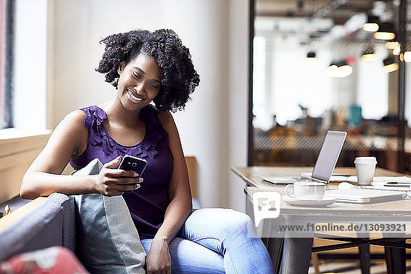 Glückliche Geschäftsfrau schaut auf Smartphone  während sie in der Büro-Cafeteria sitzt