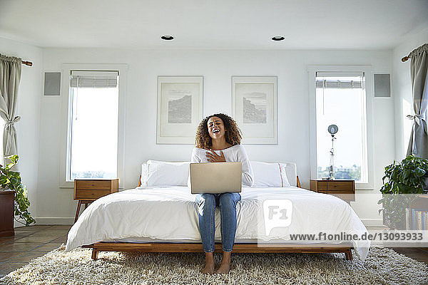 Happy woman using laptop computer while sitting on bed at home