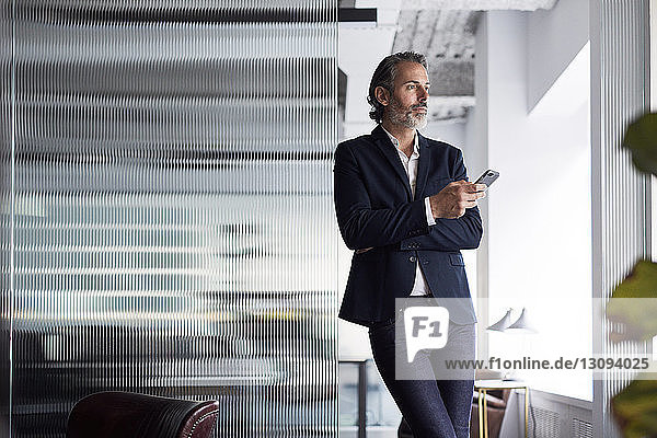 Nachdenklicher Geschäftsmann schaut weg  während er sein Smartphone im Büro an eine Glaswand hält