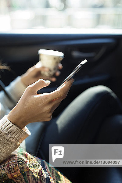 Ausgeschnittenes Bild einer Frau  die ein Smartphone benutzt  während sie im Taxi ein Einwegglas hält