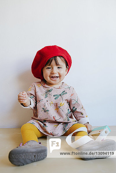 Porträt eines fröhlichen Mädchens mit rotem Hut  das zu Hause auf dem Boden an der Wand sitzt