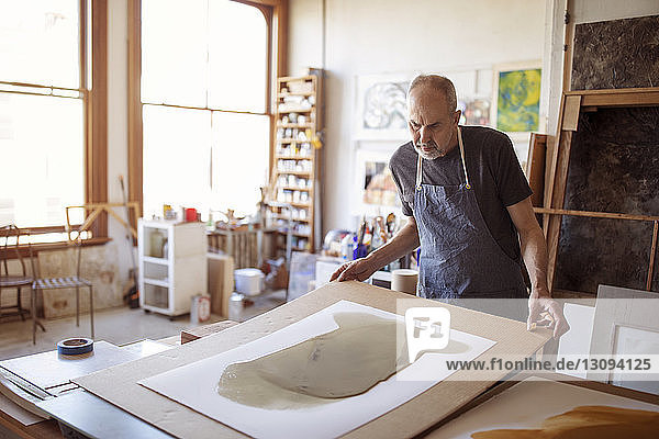 Male senior artist looking painting at table in workshop