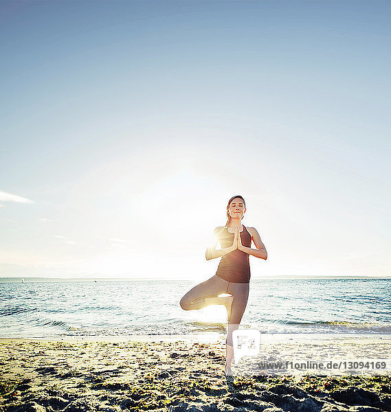 Frau meditiert in Baum-Pose am Strand gegen den Himmel