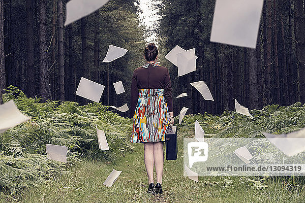 Rückansicht einer Geschäftsfrau mit Aktentasche  die inmitten herabfallender Dokumente im Wald steht