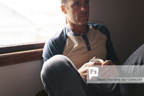 Thoughtful man with coffee cup sitting by window at home