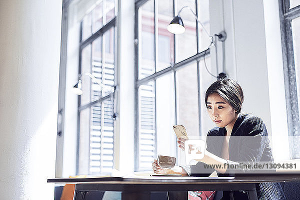 Geschäftsfrau schaut auf ein Smartphone  während sie in der Büro-Cafeteria an den Fenstern sitzt