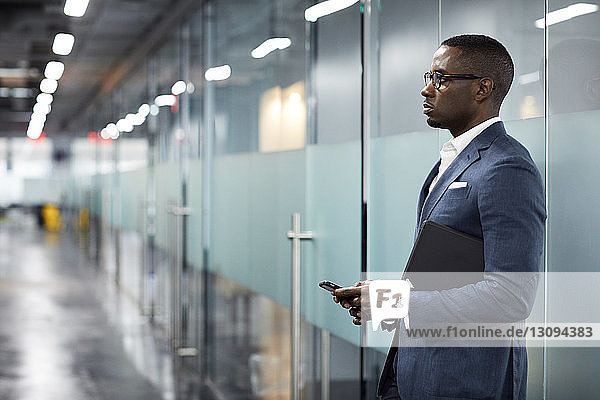 Nachdenklicher Geschäftsmann hält Smartphone in der Hand  während er in der Lobby im Büro steht
