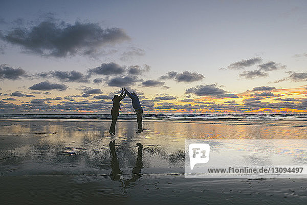 Seitenansicht eines Paares  das in Long Beach bei Sonnenuntergang vor bewölktem Himmel gemeinsam springt